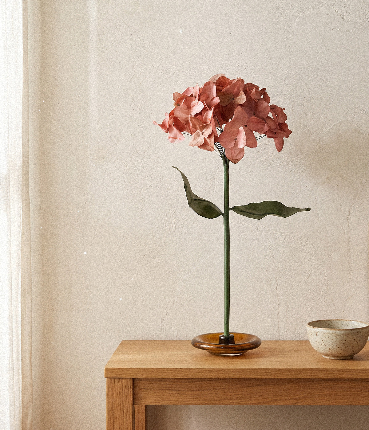 Autumn paper hydrangea on a glass stand displayed on a wood console beside a ceramic bowl.