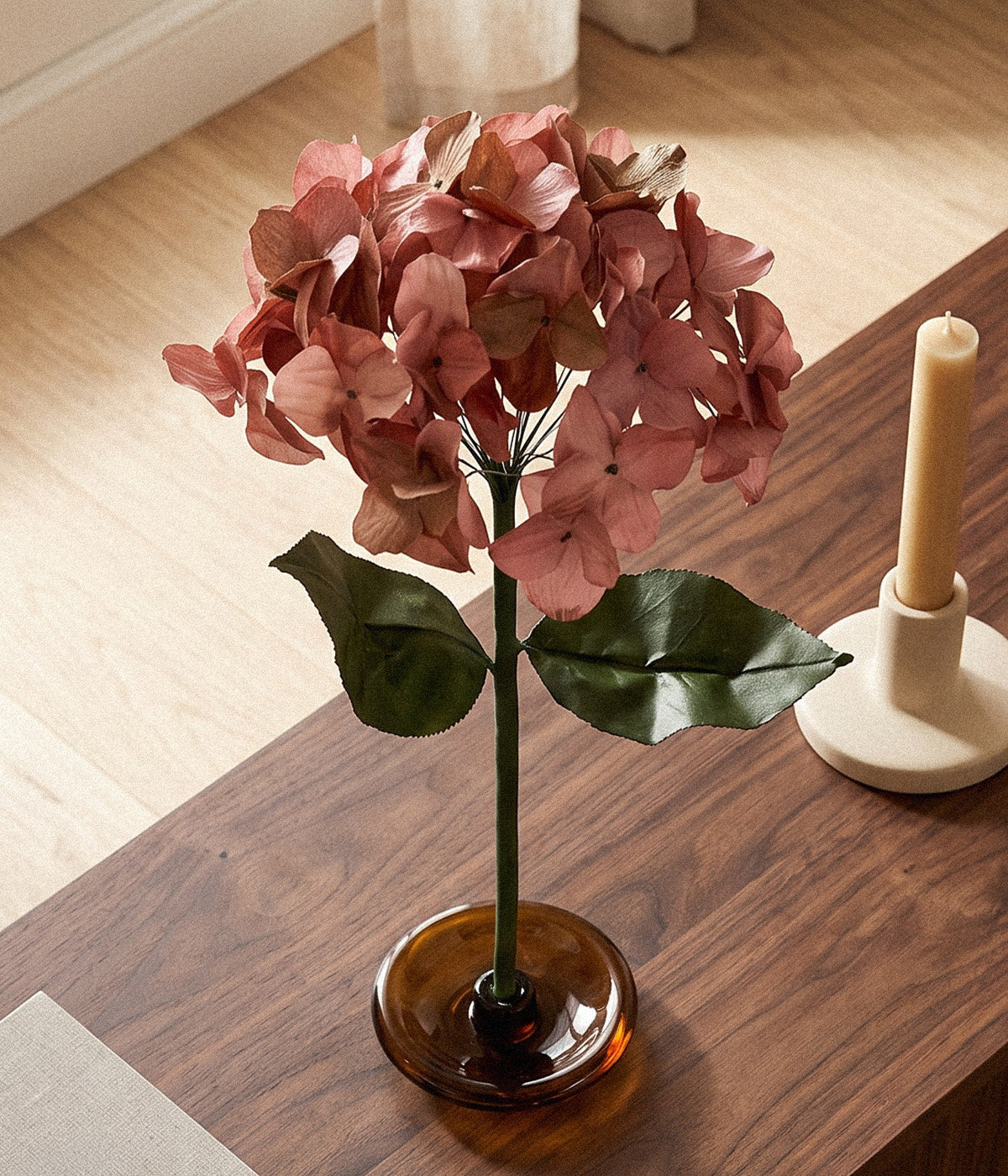 Autumn paper hydrangea with faded rose florets displayed on a glass stand beside a candle.