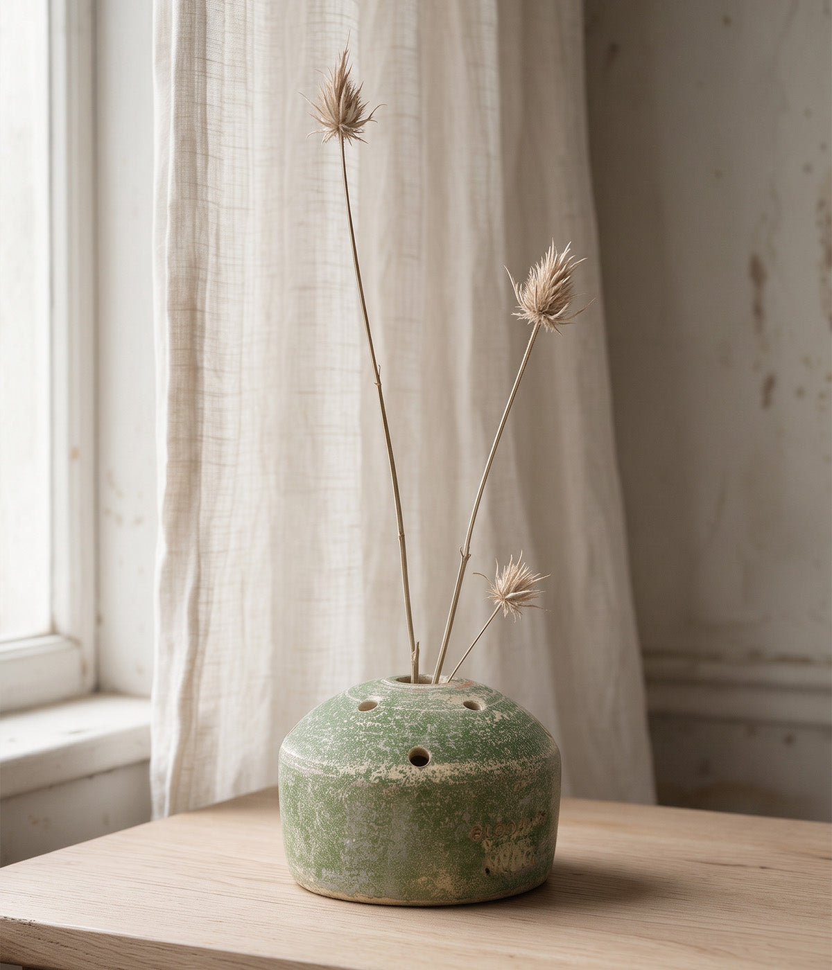 Garden Sage terracotta flower frog vase with tall dried stems displayed on a wooden table beside a softly lit window