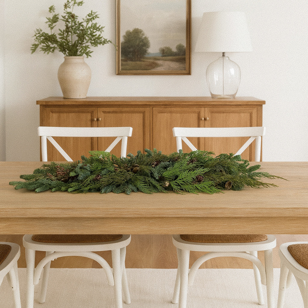 Clara juniper garland with fresh evergreens, blue berries, pine cones, and antique-style bells across a wood dining table
