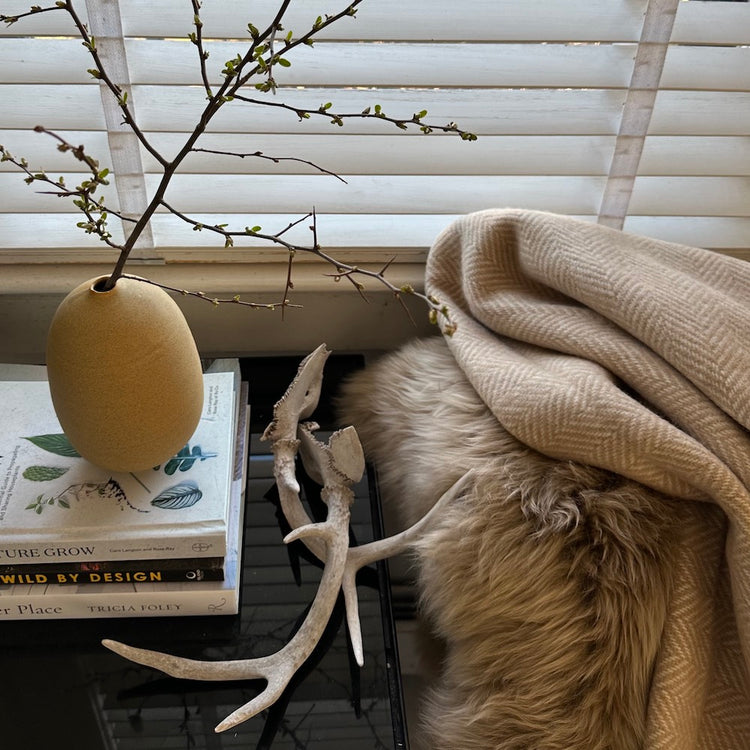 A BUTTERSCOTCH COLORED PELT WITH A HERRINGBONE THROW ON THE EDGE OF A CHAIR NEXT TO A JUDY JACKSON MUSTARD VASE  WITH A BRANCH