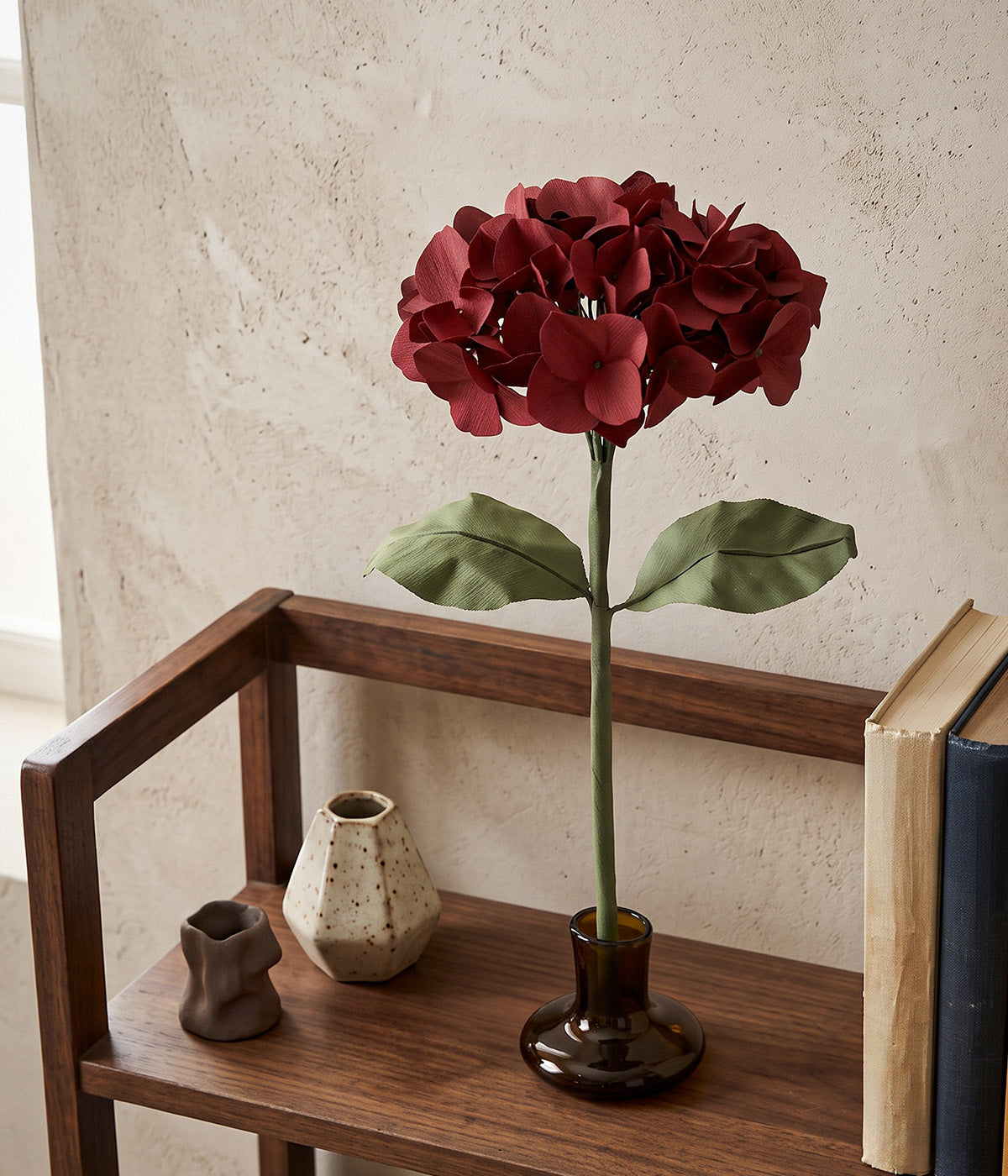 Nightfall paper hydrangea on a glass stand styled on a wooden shelf with ceramic vessels and books