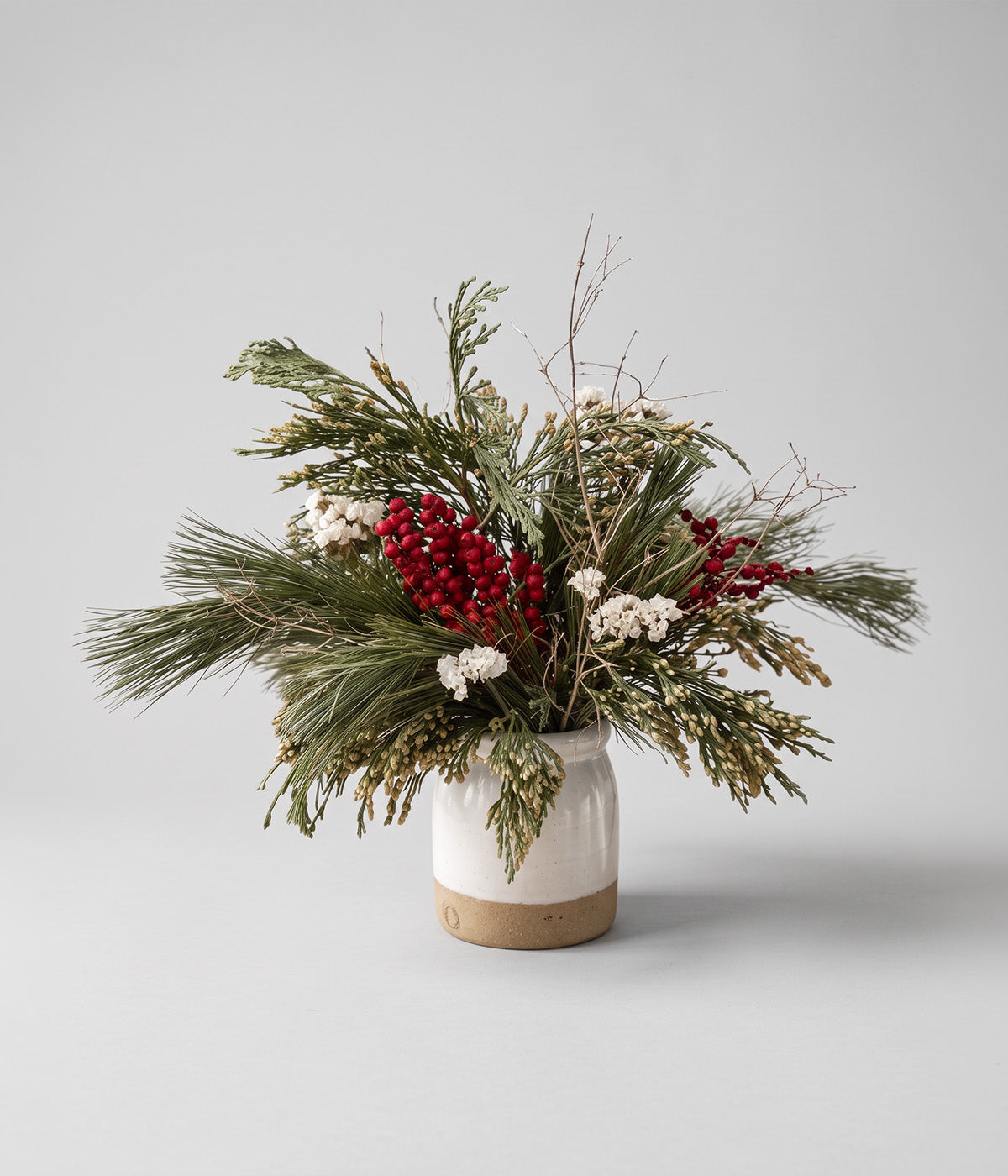 Noelle evergreen bouquet with fresh cedar, white pine, dried red canella berries, white statice, and gilded twigs in a ceramic vase
