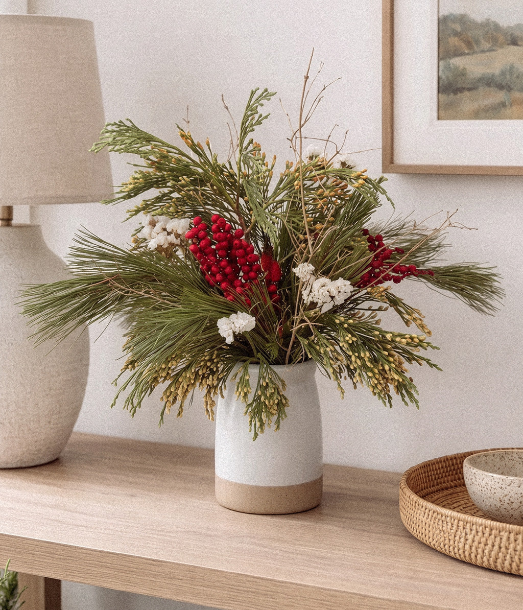 Noelle evergreen bouquet with red berries and gilded twigs arranged in a white vase on a light wood console
