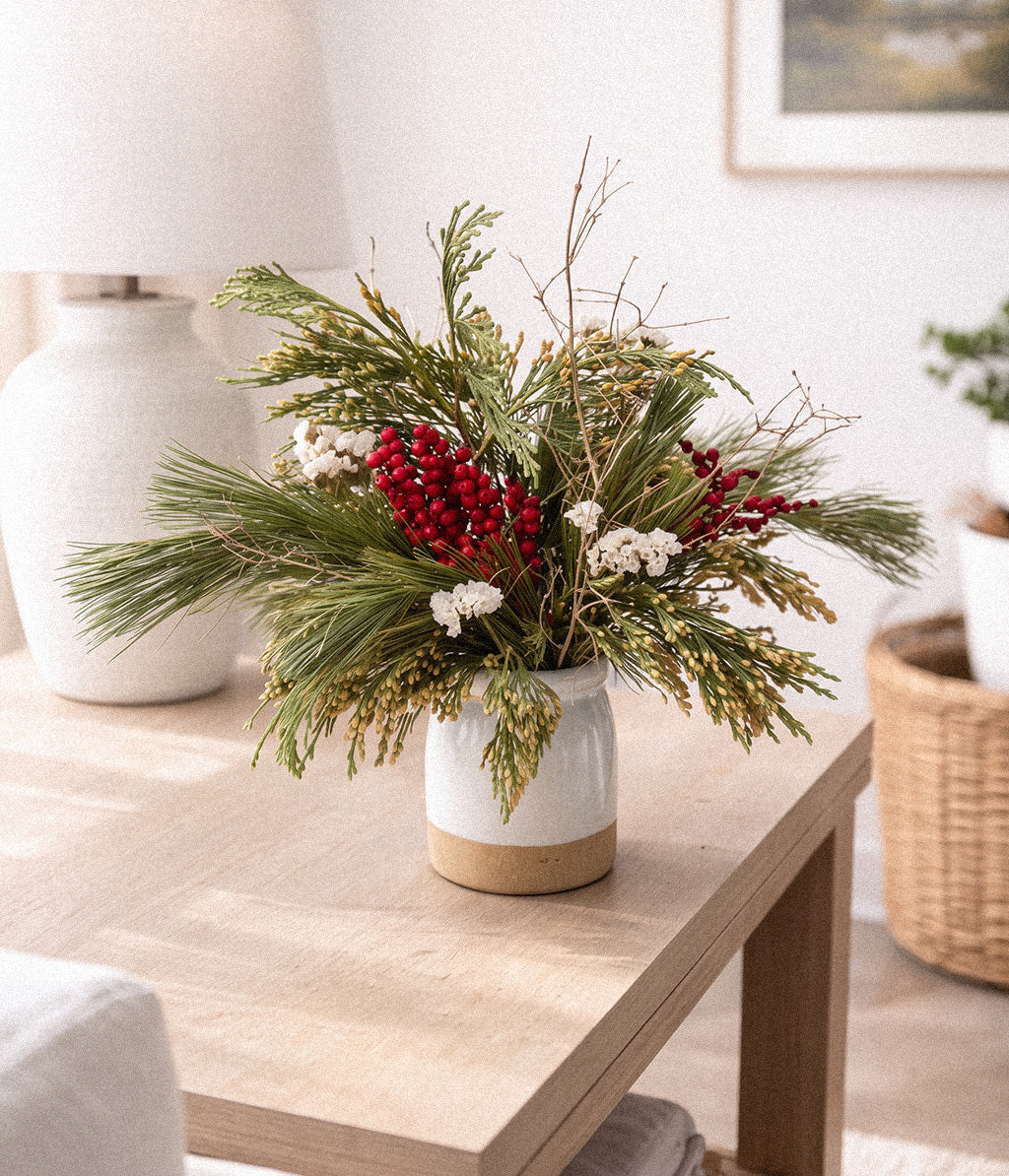 Noelle evergreen bouquet with red berries, white flowers, pine branches, and gilded twigs arranged in a white ceramic vase on a light wood side table.