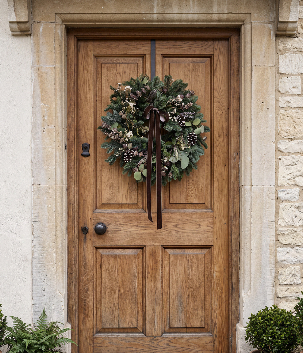 Sylvie fresh lavender and eucalyptus wreath with pine cones, white flowers, and brown velvet ribbon hanging on a wooden front door.