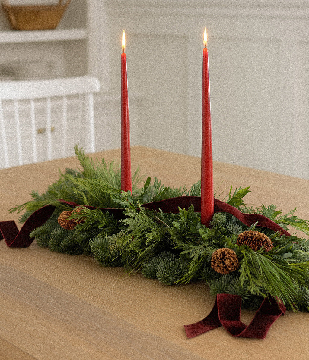 Close-up of fresh evergreen mantel runner with spruce pine cones, burgundy velvet, and red taper candles

