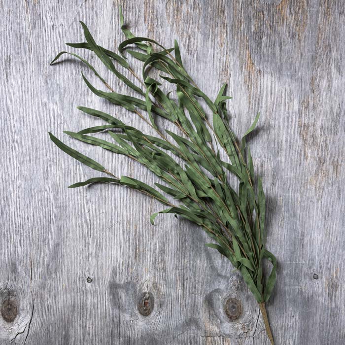 Faux Willow Branch Stem in jade with long, narrow green fabric leaves arranged on a weathered wood surface.